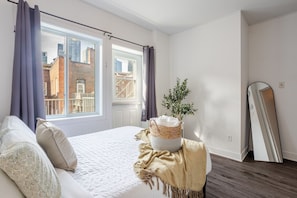A bedroom with a bed, a window with curtains, a plant, a mirror, and a view of buildings outside. - Luxurious 4 BR Suite in Griffintown (Montréal)