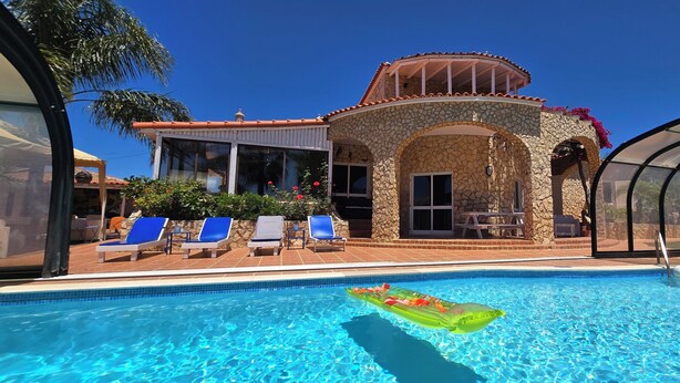 A swimming pool with a stone building in the background, a covered patio area, and a clear blue sky.
