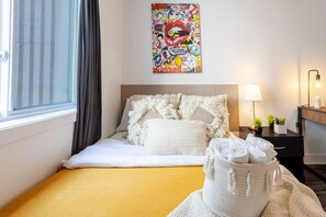 A hotel room with a bed, a window with curtains, a wall-mounted artwork, a bedside lamp, and a small plant on the nightstand.