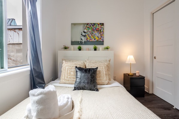 A bedroom with a bed, pillows, a bedside table with a lamp, a painting on the wall, and a window with curtains.