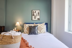 Une chambre à coucher avec un lit, des oreillers, une lampe, un tableau au mur et un panier.