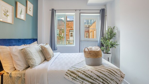 Un dormitorio con cama, ventana con cortinas, una planta e impresiones enmarcadas de motivos botánicos en la pared.