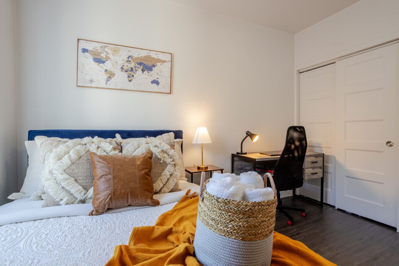Ett välordnat sovrum med en säng, kuddar, ett skrivbord, en stol, en lampa, en karta på väggen och en korg med handdukar.