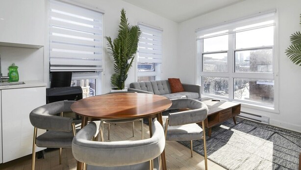 A modern living room with a round wooden table, grey upholstered chairs, a grey sofa with a red pillow, and a large window with a city view. - Contemporary 2BR Retreat in MTL`s Gay Village (Montréal)