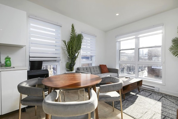 A modern living room with a round wooden table, grey upholstered chairs, a grey sofa with a red pillow, and a large window with a city view.