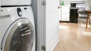 A laundry room with two front-loading washing machines, a white door, a kitchen area with a black dishwasher, and a dining table with chairs. - Contemporary 2BR Retreat in MTL`s Gay Village (Montréal)