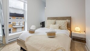 Een slaapkamer met een groot raam, een bed met witte beddengoed, twee nachtkastjes met manden, een bedlampje en een houten hoofdbord.
