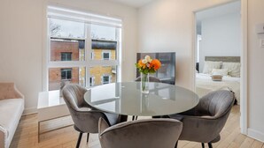 Een moderne kamer met een glazen tafel, grijze stoelen, een bed met witte beddengoed en een televisie aan de muur.