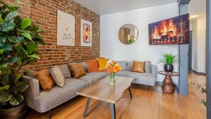A living room with a brick wall, a grey sofa, a wooden coffee table, and a flat-screen TV. - Prime Location | Charming 4BR in Downtown MTL (Montréal)