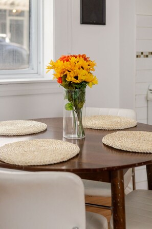 Uma mesa de jantar com um vaso de vidro transparente com flores amarelas e laranjas, rodeada por jogos americanos trançados.