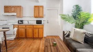 A compact kitchen with wooden cabinets, a microwave, and a stove. There's a dining area with a chair and a table, and a grey sofa with cushions. A large green plant is in the corner.