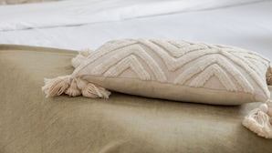A bed with a beige bedspread and a decorative pillow with tassels.