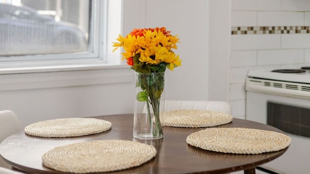 A dining table with four woven placemats and a clear vase with yellow flowers.