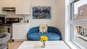 A modern kitchen with white cabinets, a black stove, and a microwave. There's a blue sofa with a decorative pillow and a vase of yellow flowers on a white table. A large abstract painting hangs above the sofa.