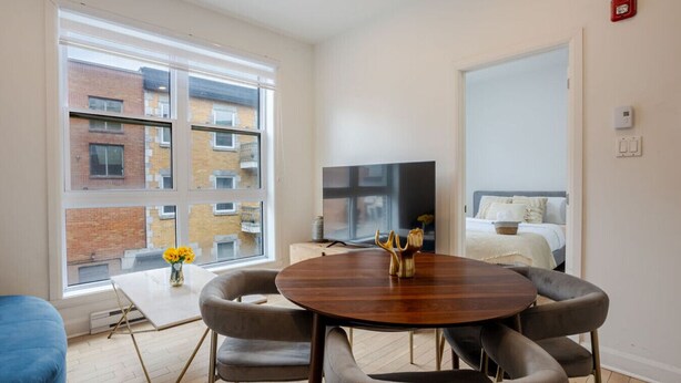 A modern living room with a round wooden table, grey upholstered chairs, a flat-screen TV, and a large window with a view of a brick building.