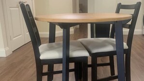 Uma mesa de jantar com quatro cadeiras, um quadro pendurado na parede e uma porta ao fundo.