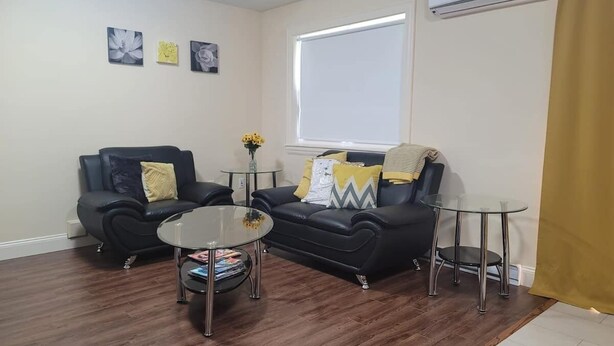 Sala de estar com sofá, duas poltronas, mesa de centro de vidro e uma mesinha lateral. Há uma janela com persiana branca, um aparelho de ar condicionado na parede e uma cortina amarela parcialmente visível.