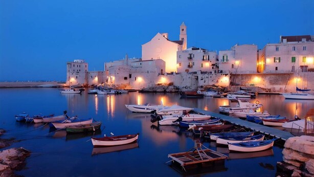 Une ville côtière au crépuscule, avec des bateaux amarrés le long du port, un bâtiment imposant doté d’une tour, et une mer calme qui reflète les lumières.