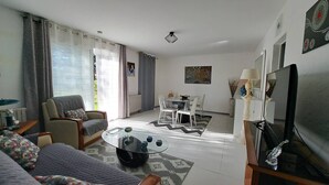 A living room with a sofa, armchair, coffee table, dining table, and chairs. There is a television and a lamp on a stand.