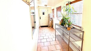 Un couloir avec un sol carrelé, une porte avec une fenêtre, une étagère en métal ornée de plantes et un porte-manteau fixé au mur.