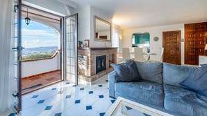 Un salon avec une cheminée, un canapé, un coin repas et une vue sur l’extérieur par une porte ouverte.