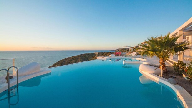 A infinity pool with a view of the sea, a palm tree, and a white building.