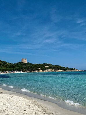 Een strand met helder turquoise water, een zandstrand en een kasteelachtige constructie op een heuvel.