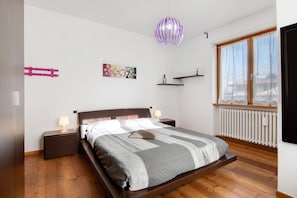 Un dormitorio con cama, mesitas de noche, lámpara colgante, radiador y ventana con cortinas.