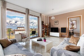 Un salón con sofá, mesa de centro, televisor, estantería y un balcón con vistas.
