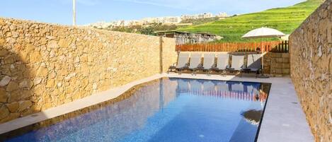 A swimming pool with stone walls, a view of a hillside, and a row of sun loungers.