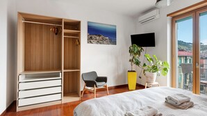 Un dormitorio con un armario de madera, una cama con colcha blanca estampada, una silla, una planta en maceta y vistas a edificios a través de un ventanal grande.