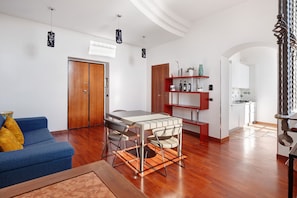 Una sala moderna con un sofá azul, una mesa de comedor y sillas, un armario de madera, y un área de cocina visible a través de un arco.