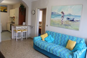 Una sala de estar con un sofá azul, un cuadro de una mujer junto al mar, una cocina con armarios blancos y una zona de comedor con sillas blancas.