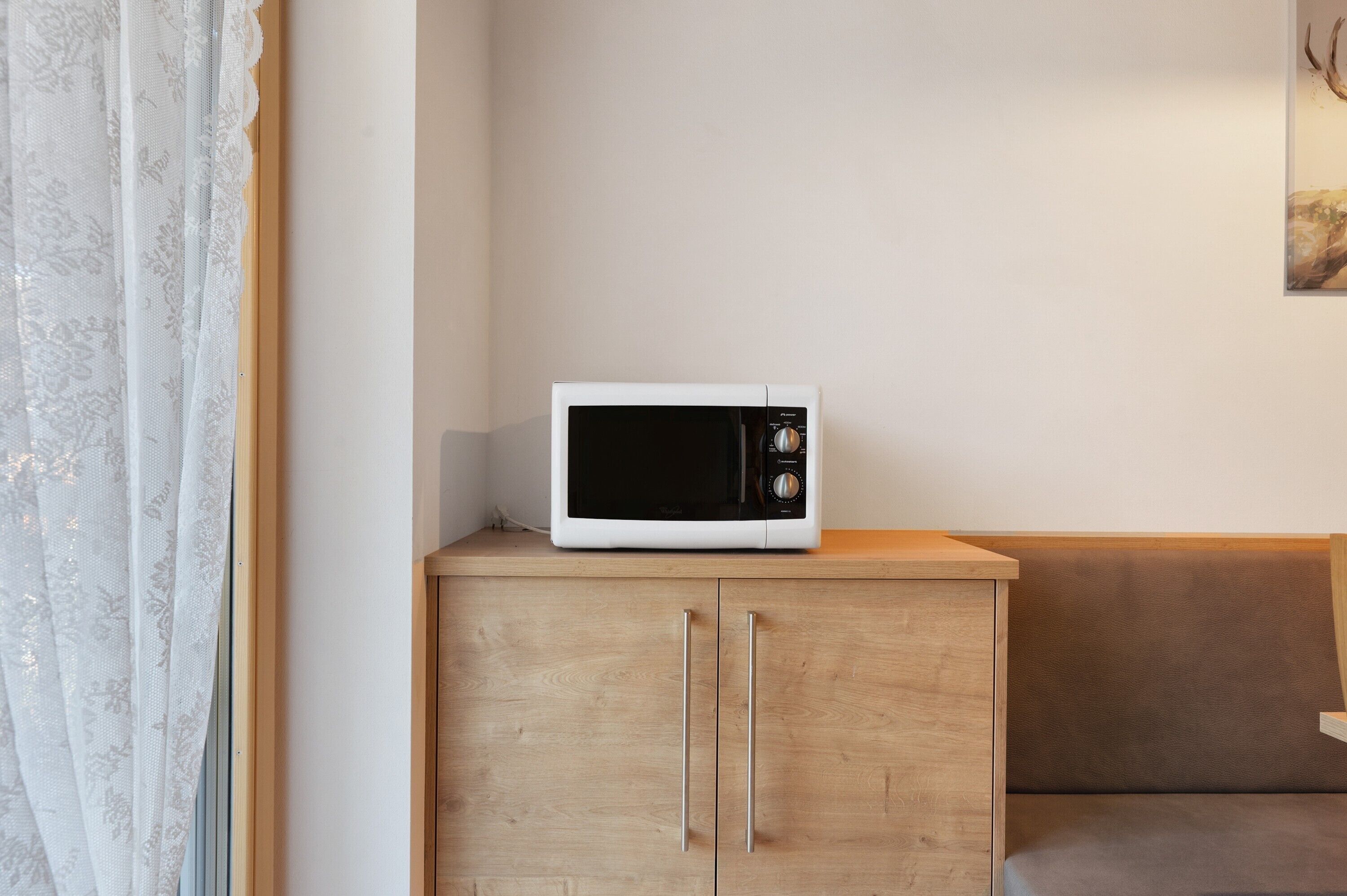 Un four à micro-ondes blanc posé sur un meuble en bois, un canapé marron et une fenêtre avec des rideaux légers.