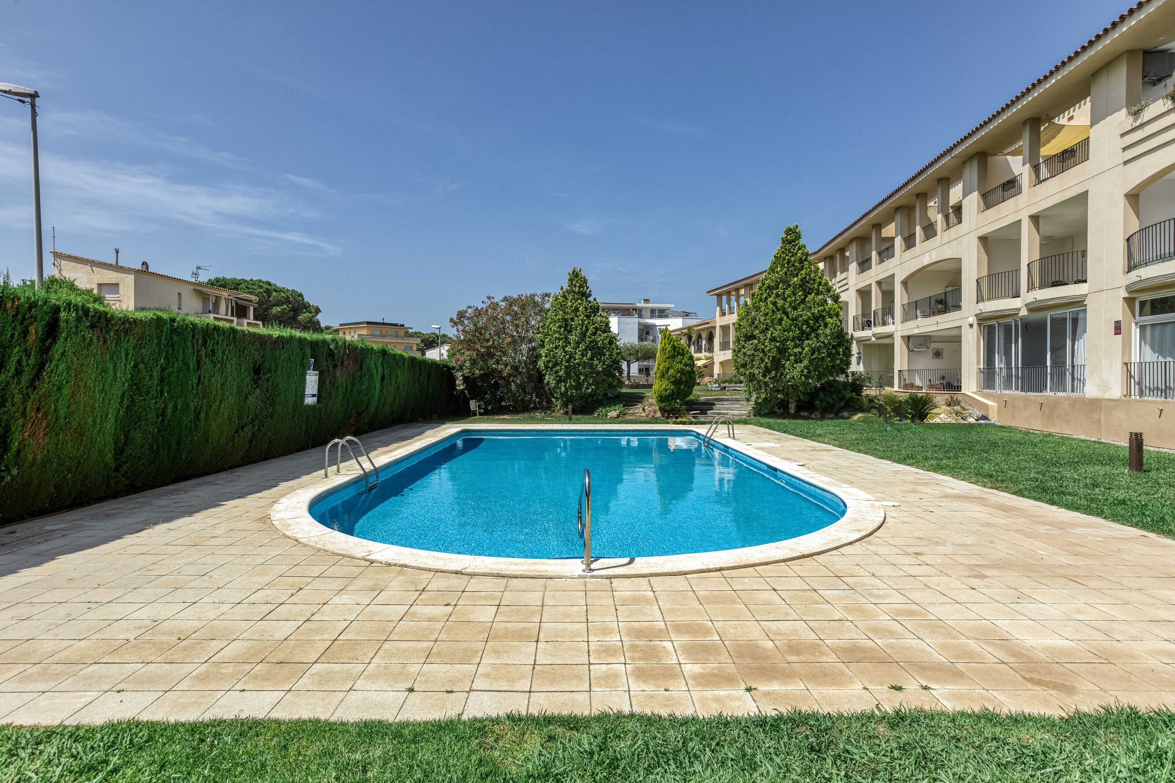 A swimming pool with a tiled deck, surrounded by a hedge and a building with balconies.