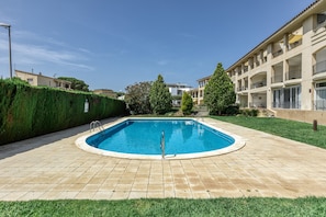 A swimming pool with a tiled deck, surrounded by a hedge and a building with balconies.