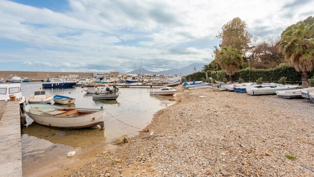 Un port de plaisance où des bateaux sont amarrés le long d’une rive de galets, sous un ciel dégagé, avec des montagnes qui se dessinent à l’horizon.