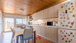 Une cuisine avec des meubles en bois, un réfrigérateur orné d’aimants, une table à manger avec des chaises, et une vue à travers une porte vitrée.