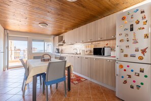 Une cuisine avec des meubles en bois, un réfrigérateur orné d’aimants, une table à manger avec des chaises, et une vue à travers une porte vitrée.