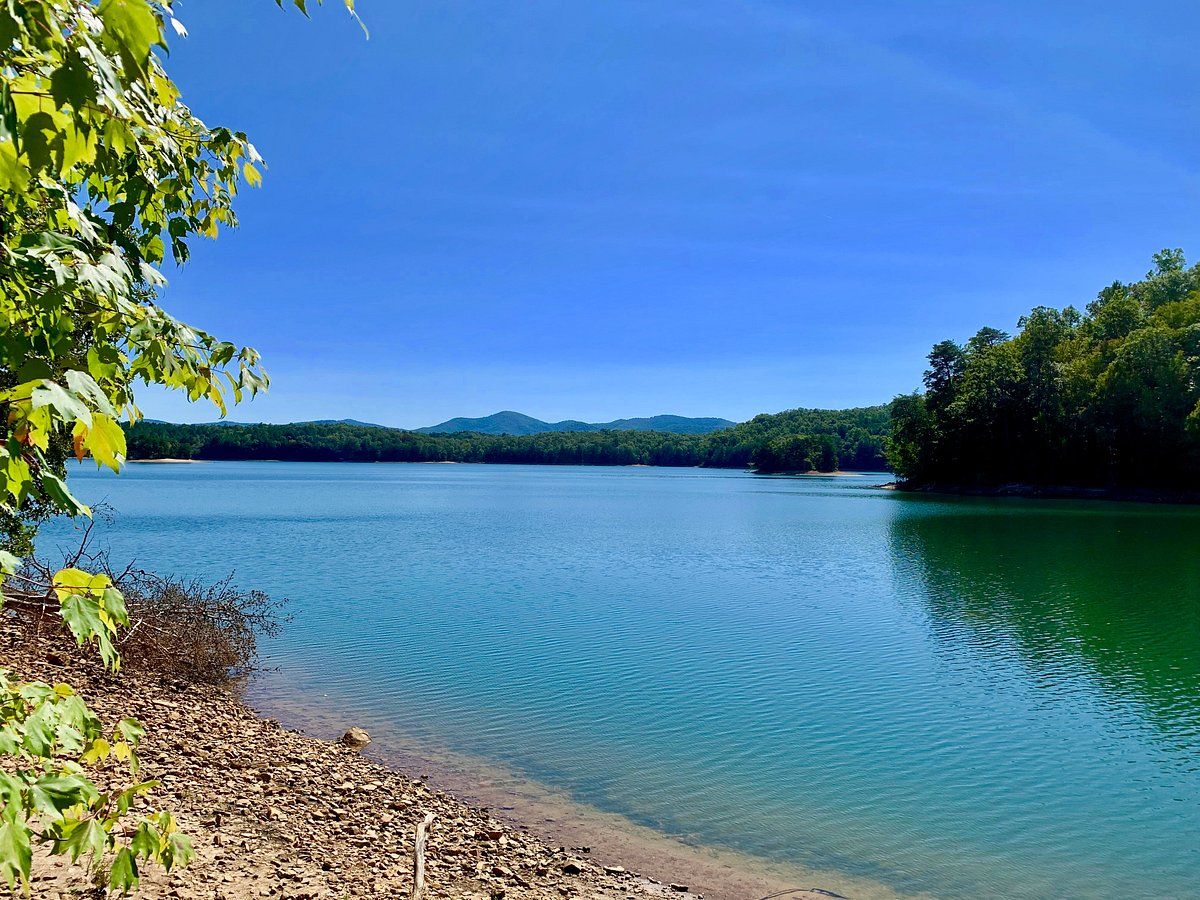 A calm lake with clear water, surrounded by dense green forest and distant hills.