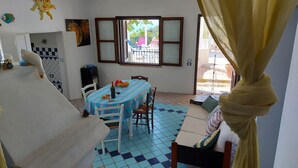 Ein Zimmer mit Esstisch, Stühlen, einer Couch, einem Fenster und Blick ins Freie.