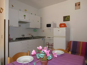 A small kitchen with white cabinets, a microwave, and a refrigerator. There is a dining table with chairs and a vase of flowers.