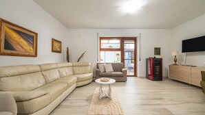 A living room with a beige sofa, a small round table, and a television on the wall. - Casa Vacanze 'EliFra' with Private Terrace, Garden, and Balcony (San Giovanni Suergiu)