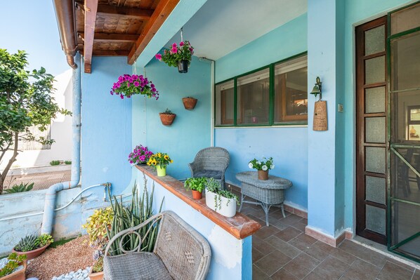 Pared azul exterior con cestas de flores colgantes, una silla de mimbre y una mesita.