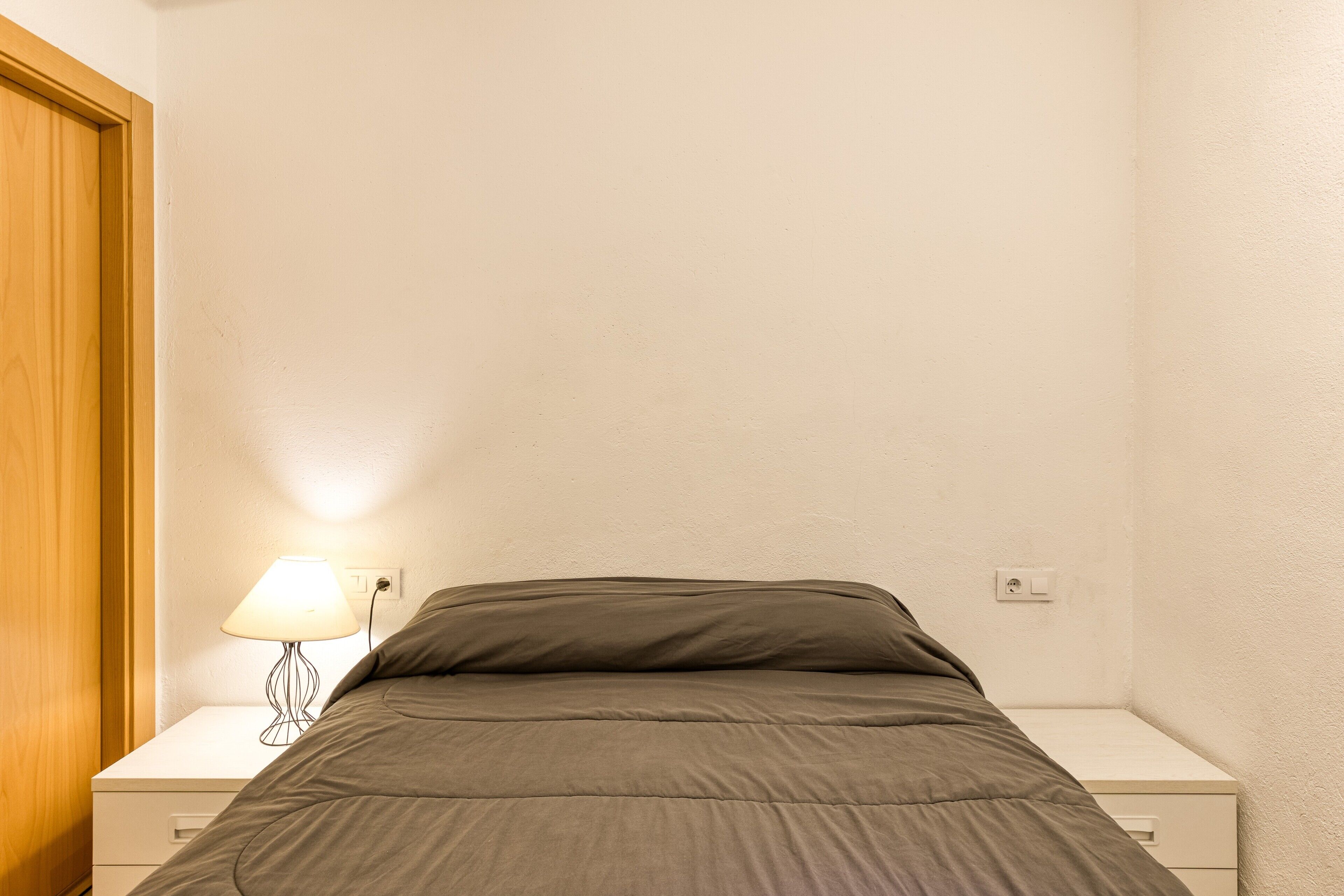 A single bed with a grey blanket, a bedside table with a lamp, and a wooden door.