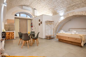A room with a stone ceiling, a bed with a wooden headboard, a dining area with chairs, a small kitchen with a wooden cabinet, and a bathroom with a towel rack. - Studio 'La Pizzicata' with Wi-Fi and Air Conditioning (Ostuni)