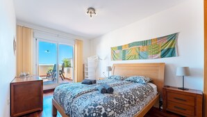Ein modernes Schlafzimmer mit einem großen Bett, einem Nachttisch aus Holz, einer Nachttischlampe, Blick ins Freie und einem farbenfrohen Wandteppich.