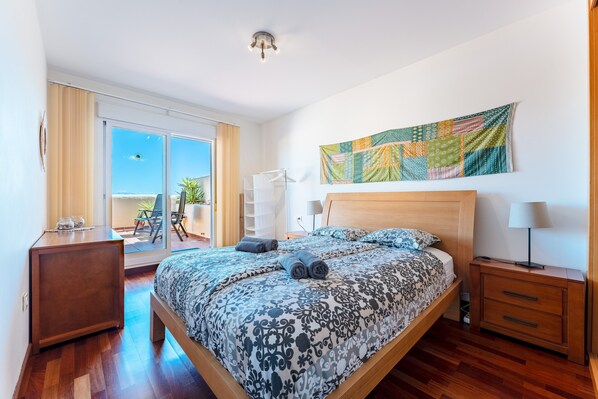 Une chambre moderne avec un grand lit, une table de chevet en bois, une lampe de chevet, une vue sur l’extérieur et une tapisserie colorée au mur.