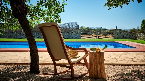 A poolside chair with a woven seat, a wooden stump with a glass of drink, and a clear view of a swimming pool.