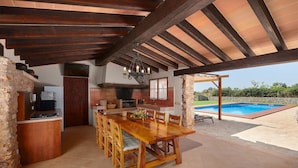 A spacious outdoor dining area with a wooden table and chairs, a stone wall, a pool, and a pergola.